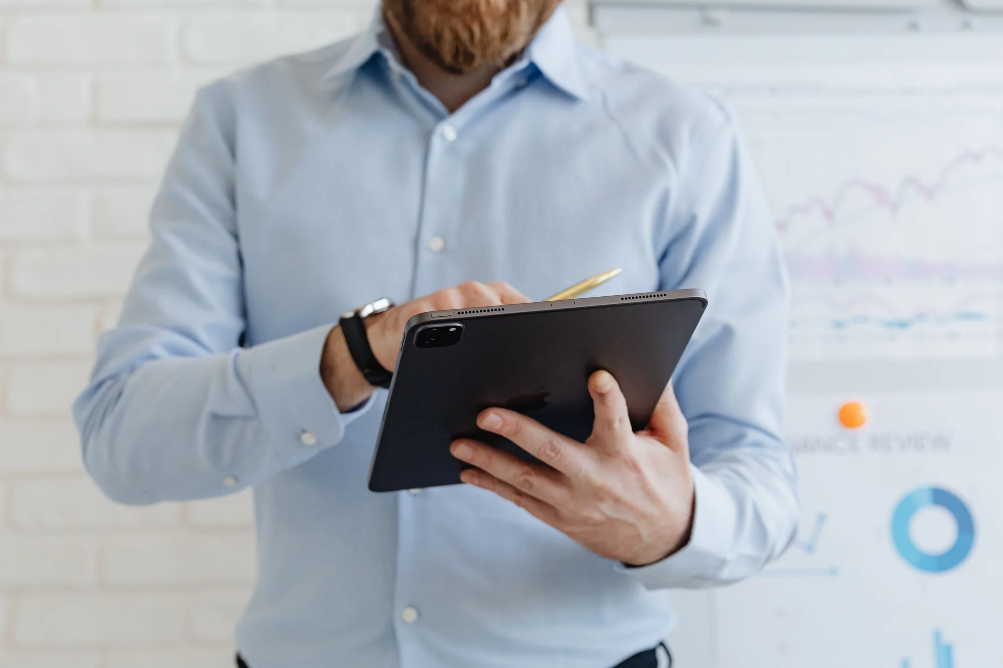 Man in a light blue shirt holding a tablet and a gold pen in front of a whiteboard with charts